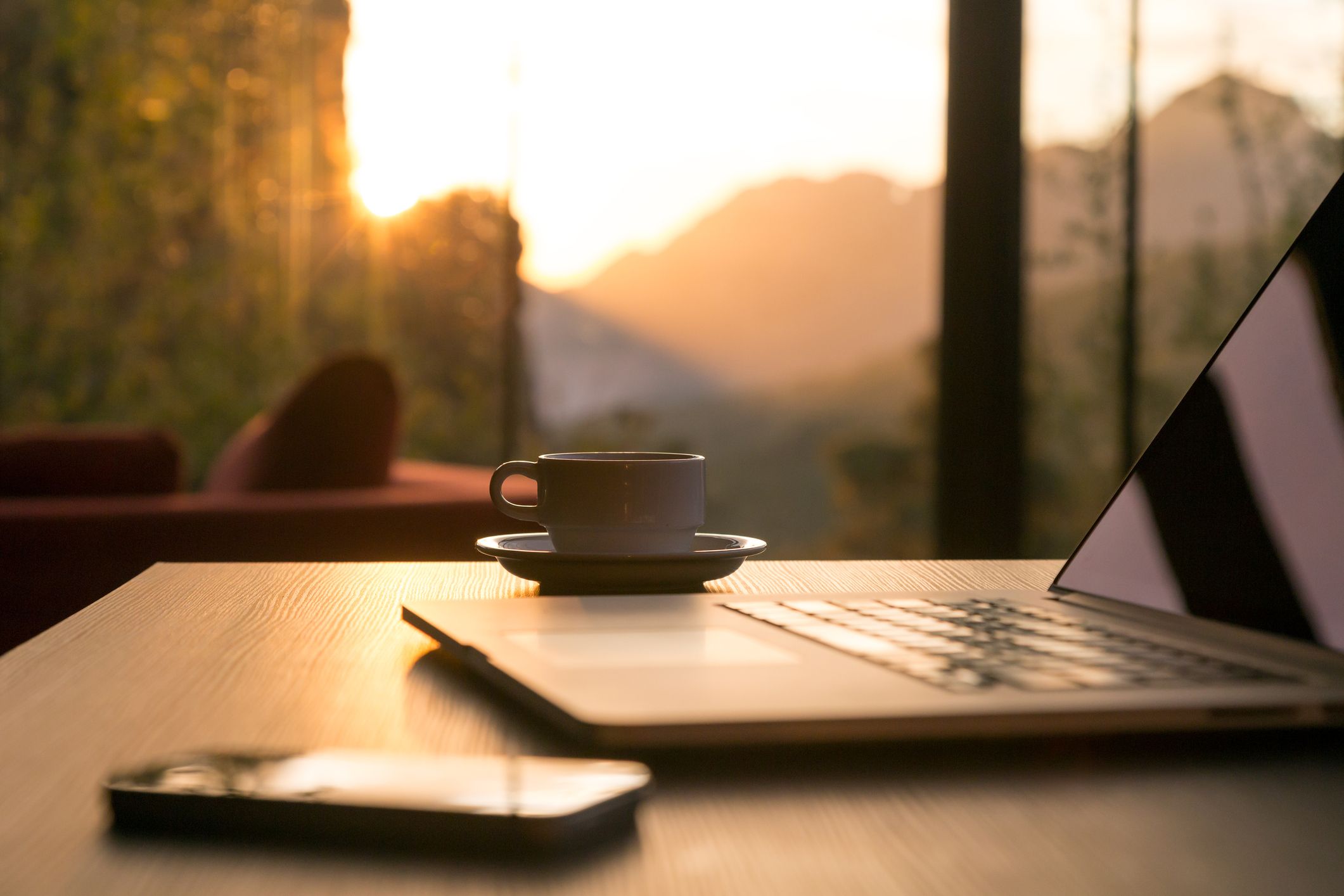 On a wooden table in a home office, a person is using a laptop while a cup of coffee and a smartphone are sitting on the table.