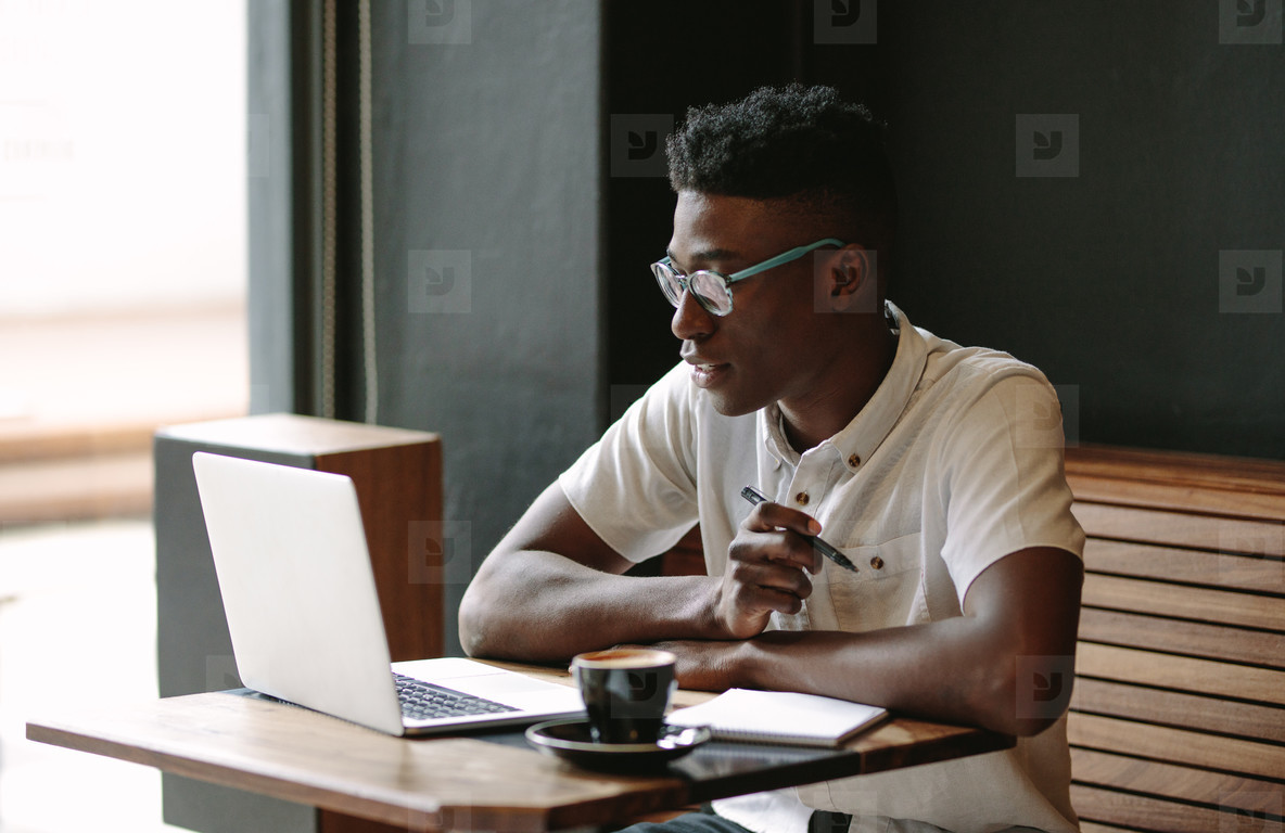 A young man wearing glasses is working on his laptop while sitting at a cafe table. He has a cup of coffee on the table and is holding a pen.
