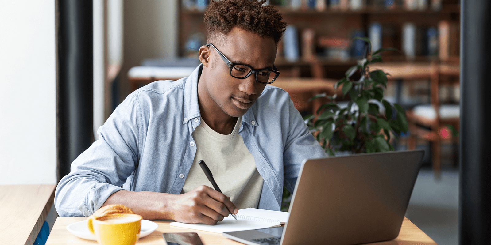 A man wearing glasses is writing in a notebook while looking at his laptop; he is using a freelance platform to find qualified freelancers for his project.