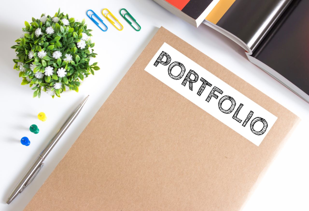 A notebook labeled 'PORTFOLIO' sits on a desk next to a pen, some books, paper clips, and a plant, illustrating the search query 'Tips to improve skills and portfolio for freelancers through online courses'.
