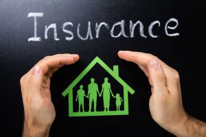 A green paper cut out of a family in a house is held up in front of a blackboard with the word 'insurance' written in white chalk, with a pair of hands cupping around the paper cut out.