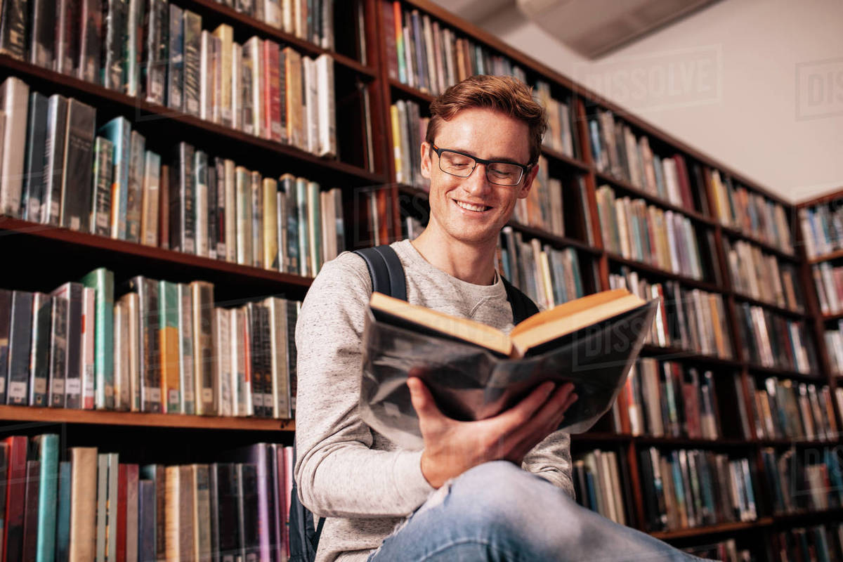 A young man wearing glasses sits on the floor of a library surrounded by bookshelves, smiling while reading an open book.