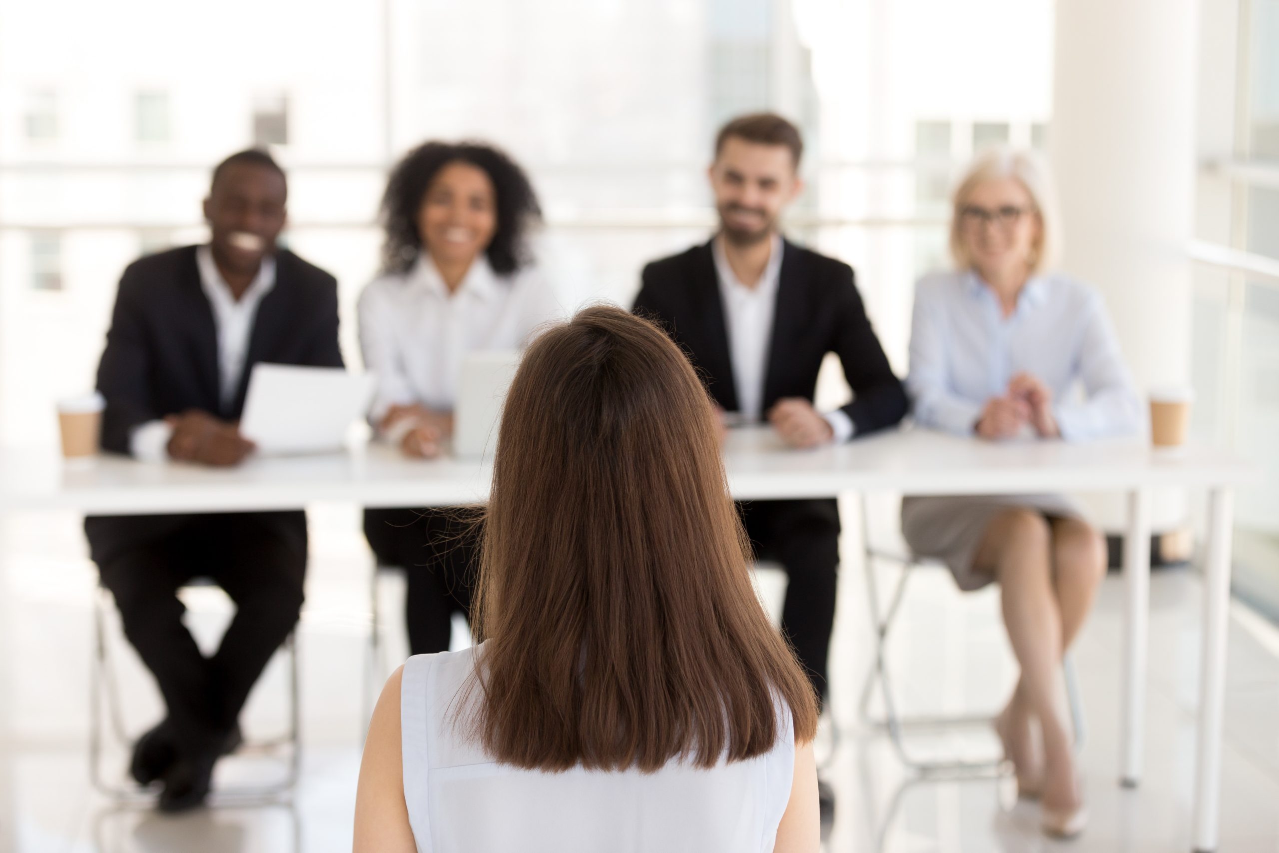 A woman sits with her back to the camera in front of a diverse hiring panel of four people.