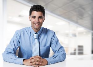 A successful businessman wearing a blue shirt and tie is sitting at his desk in a modern office and looking at the camera with a smile on his face.