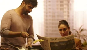 A man and woman in a kitchen, the man is cooking while the woman is reading the newspaper with the headline 'Impact of wife earning more than husband'.