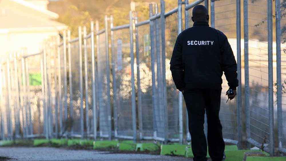 A security guard wearing a black uniform with a SECURITY patch on the back is patrolling a fenced area.
