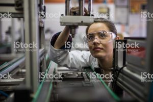 A female factory worker wearing safety glasses inspects a machine.