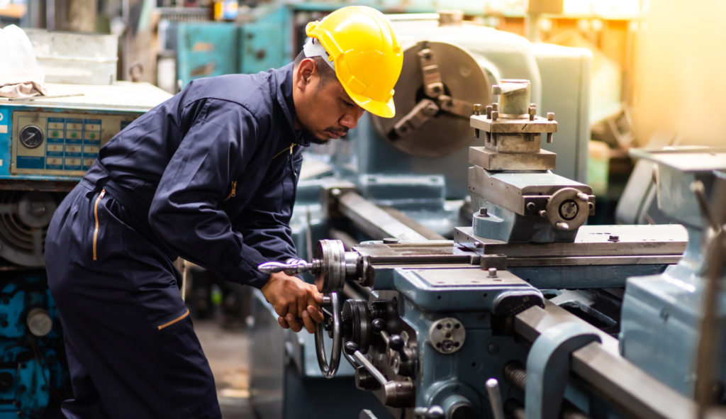 A production operator wearing a hard hat and safety glasses operates a machine in a factory.