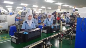 The image shows a production operator wearing a hijab and blue uniform operating a machine in a factory.