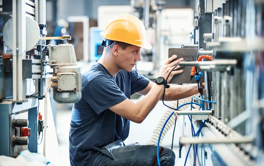 A technician wearing a yellow hard hat and blue work clothes operates a machine in a production environment.