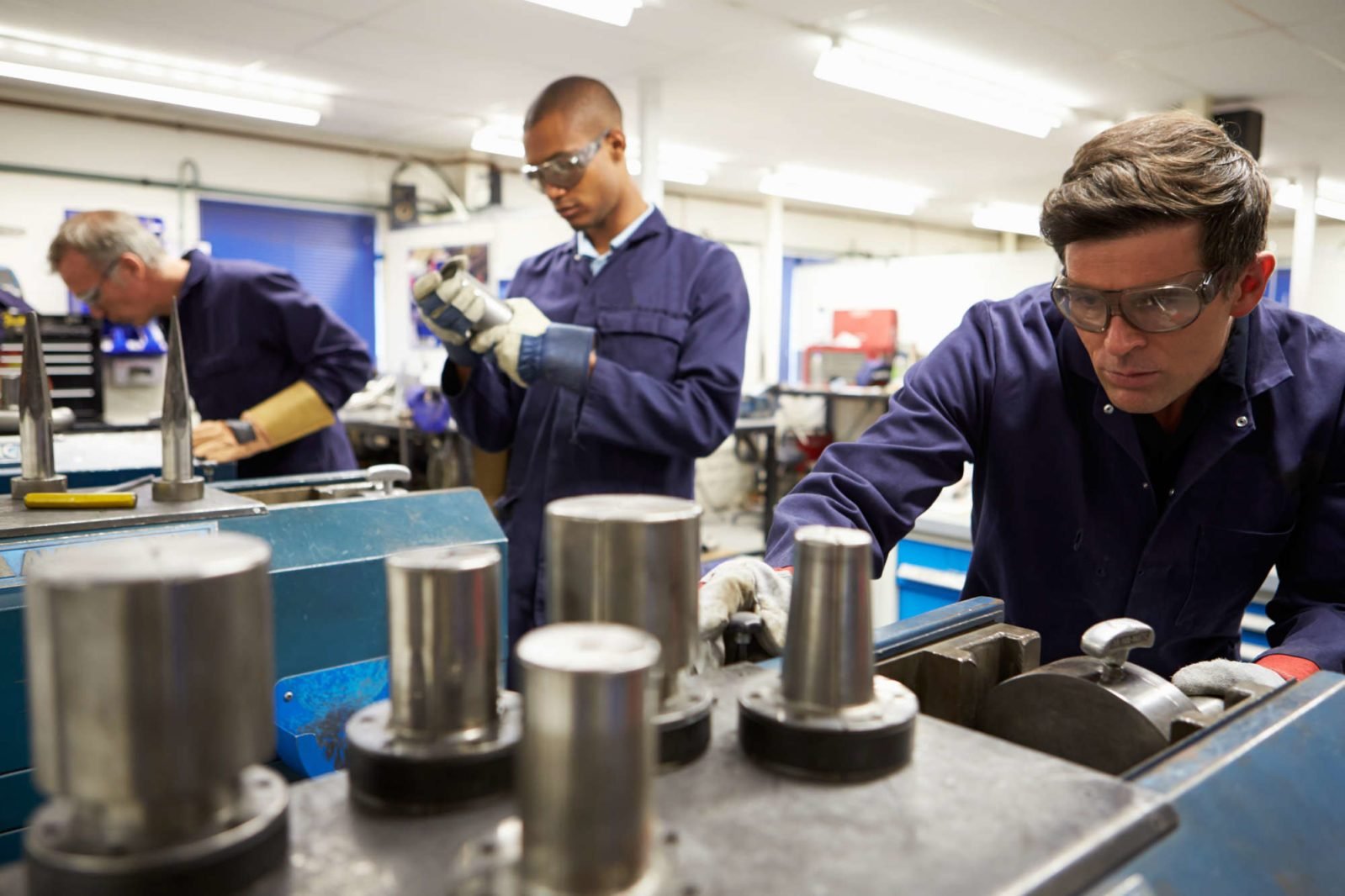 In a factory, a skilled production operator wearing safety glasses operates an industrial machine while two coworkers are working in the background.