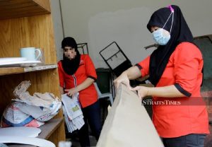 Two female supervisors wearing red shirts and face masks sort through a pile of documents and boxes in a storeroom.