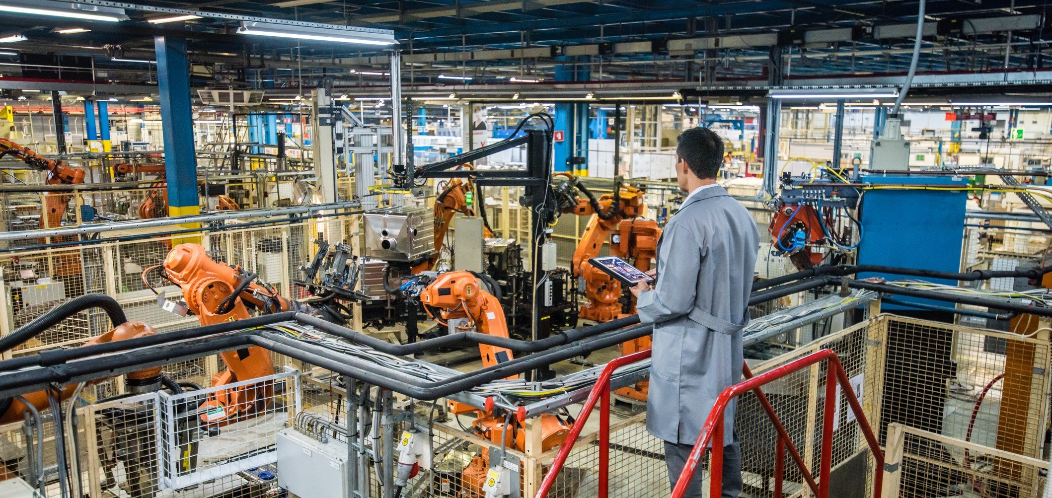 A factory supervisor in a white coat stands on a catwalk overlooking the production floor of a factory, where robotic arms are working.