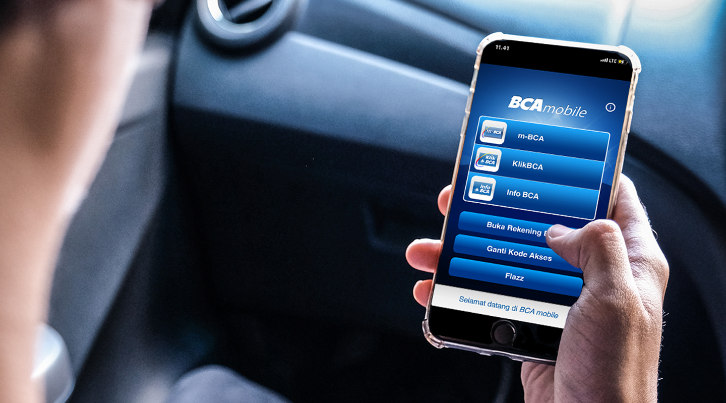 A hand holding a smartphone with the BCA Mobile Banking app pulled up, which has an option to top up an electronic toll collection card.