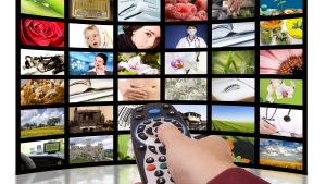 A hand holding a remote control is pointed at aの一覧 of TV channels with various programs, including Penyebab gangguan sinyal TV digital.