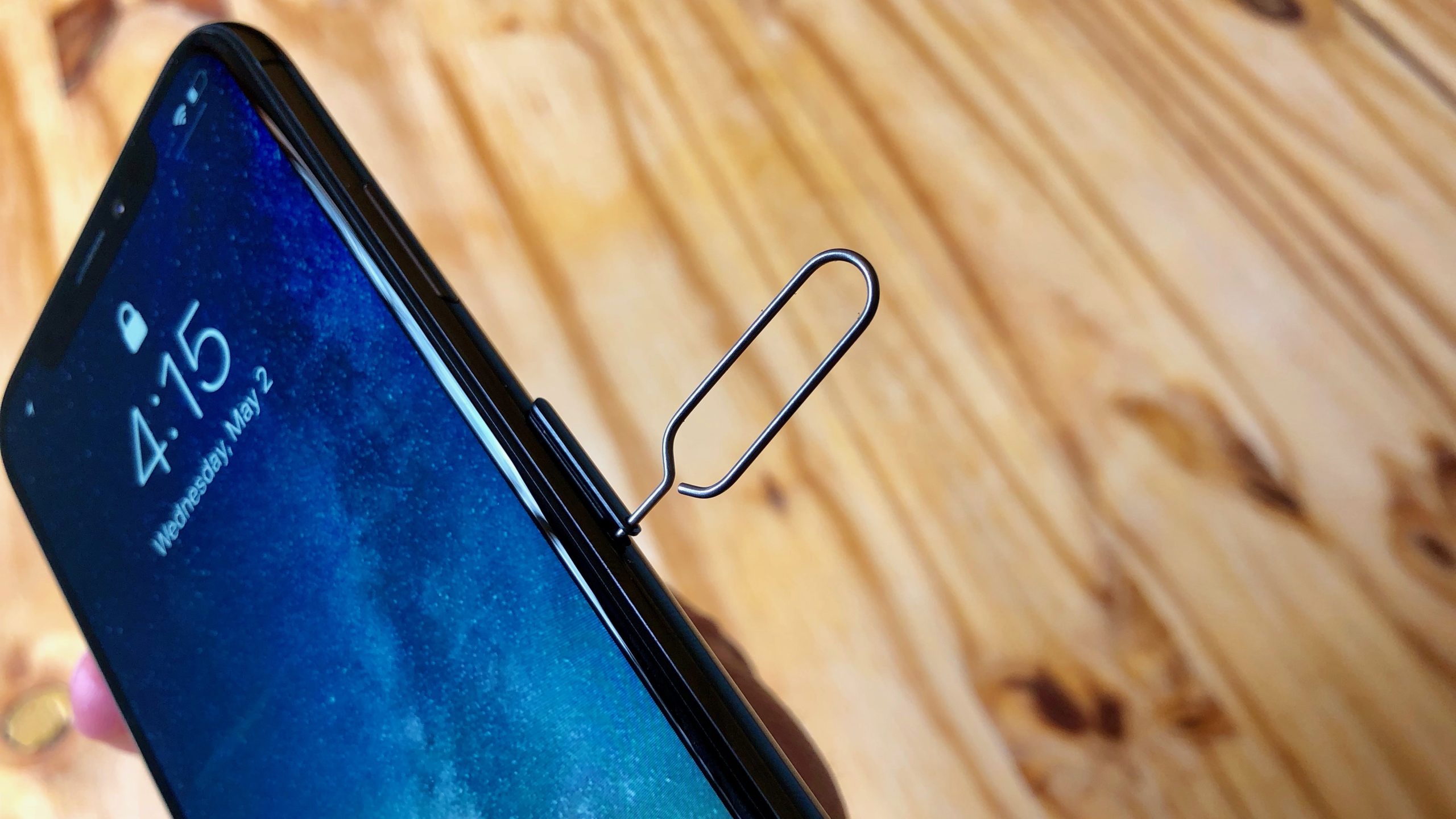A person holding an iPhone with a paperclip inserted in the SIM card slot, with a wooden background.