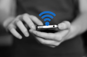 A grayscale image of a person holding a phone with a blue Wi-Fi symbol superimposed on the phone in front of an out of focus background of water and trees.