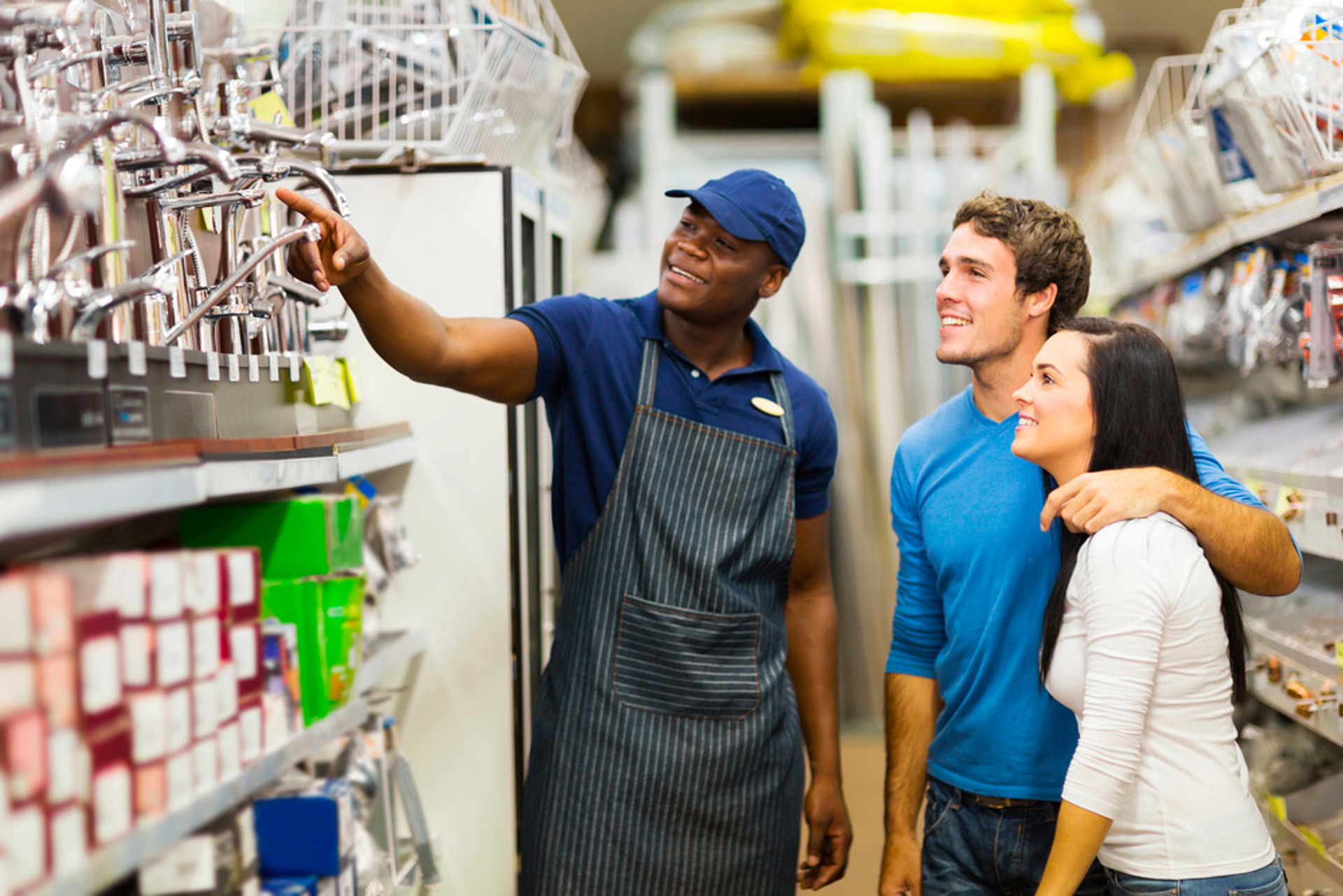 A smiling sales clerk wearing a blue apron assists a young couple in a hardware store.