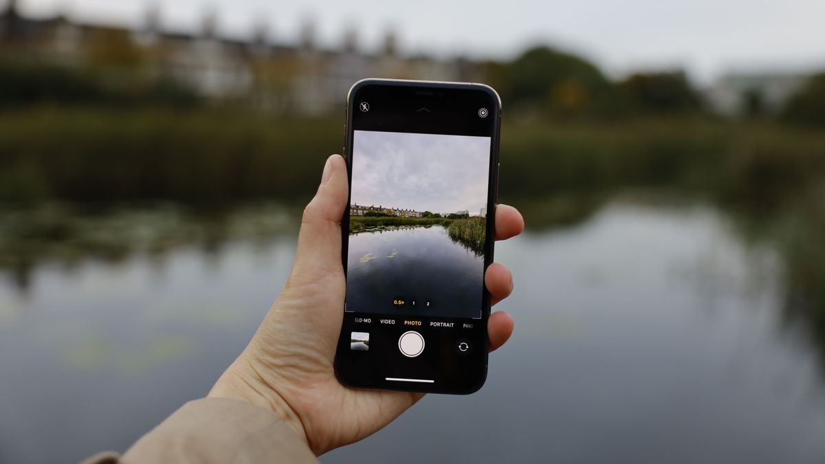 A hand holding a black smartphone taking a picture of a lake and trees in the background.