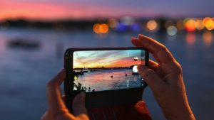 A person holds a smartphone in their hand and takes a picture of a sunset over a river.