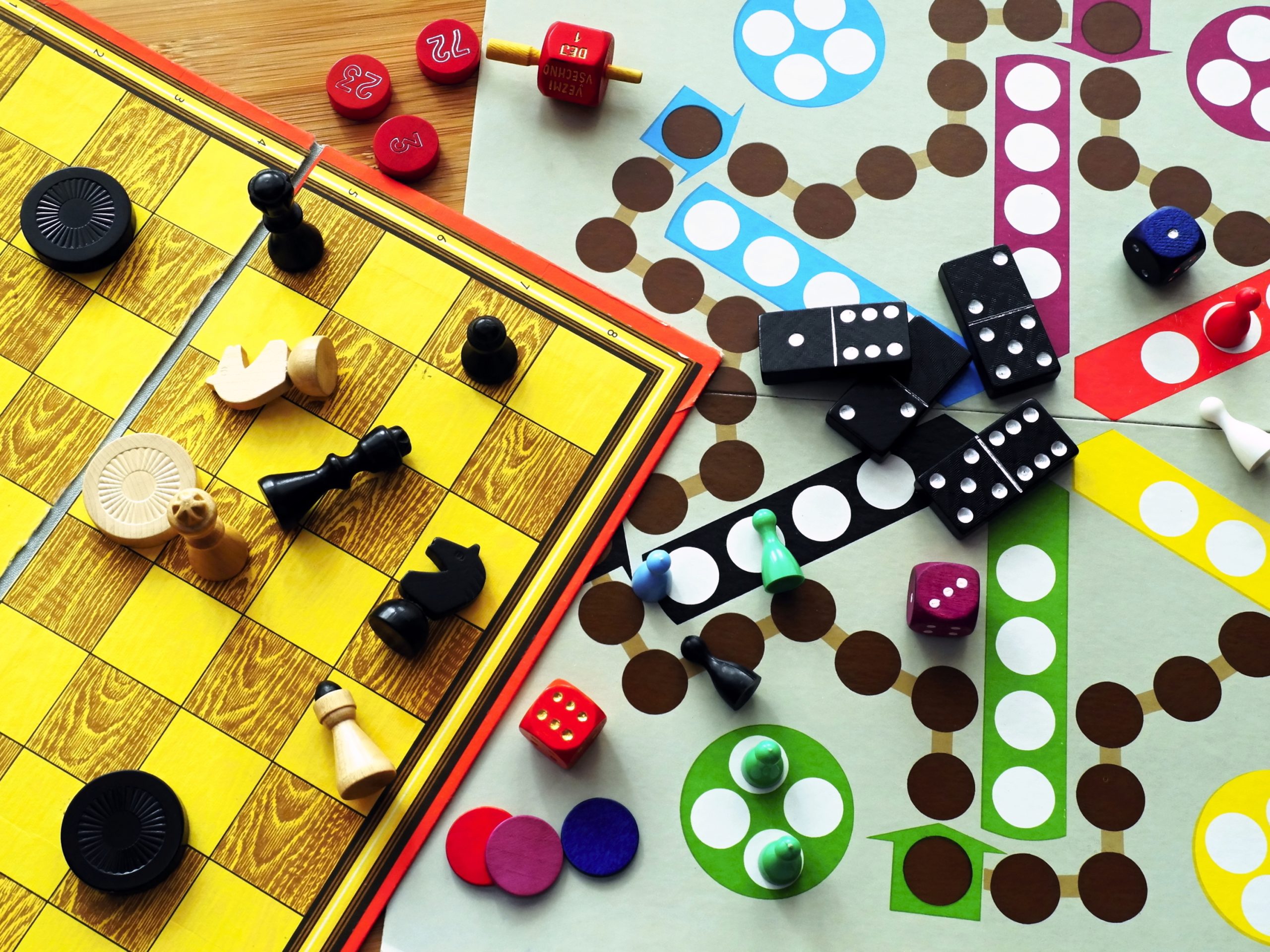 A variety of board games and blocks are arranged on a wooden table including chess, checkers, dominoes, and a spinner.