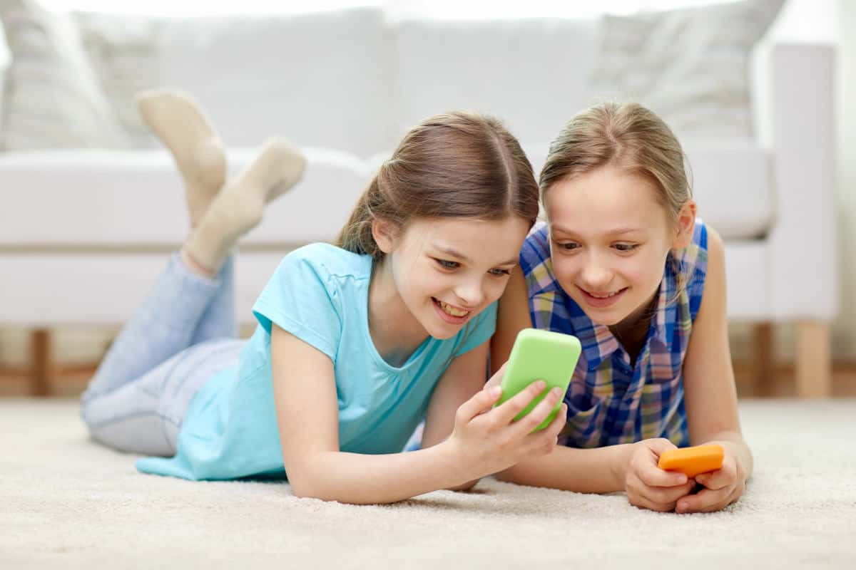 Two girls lying on the floor looking at their phones with smiles on their faces.